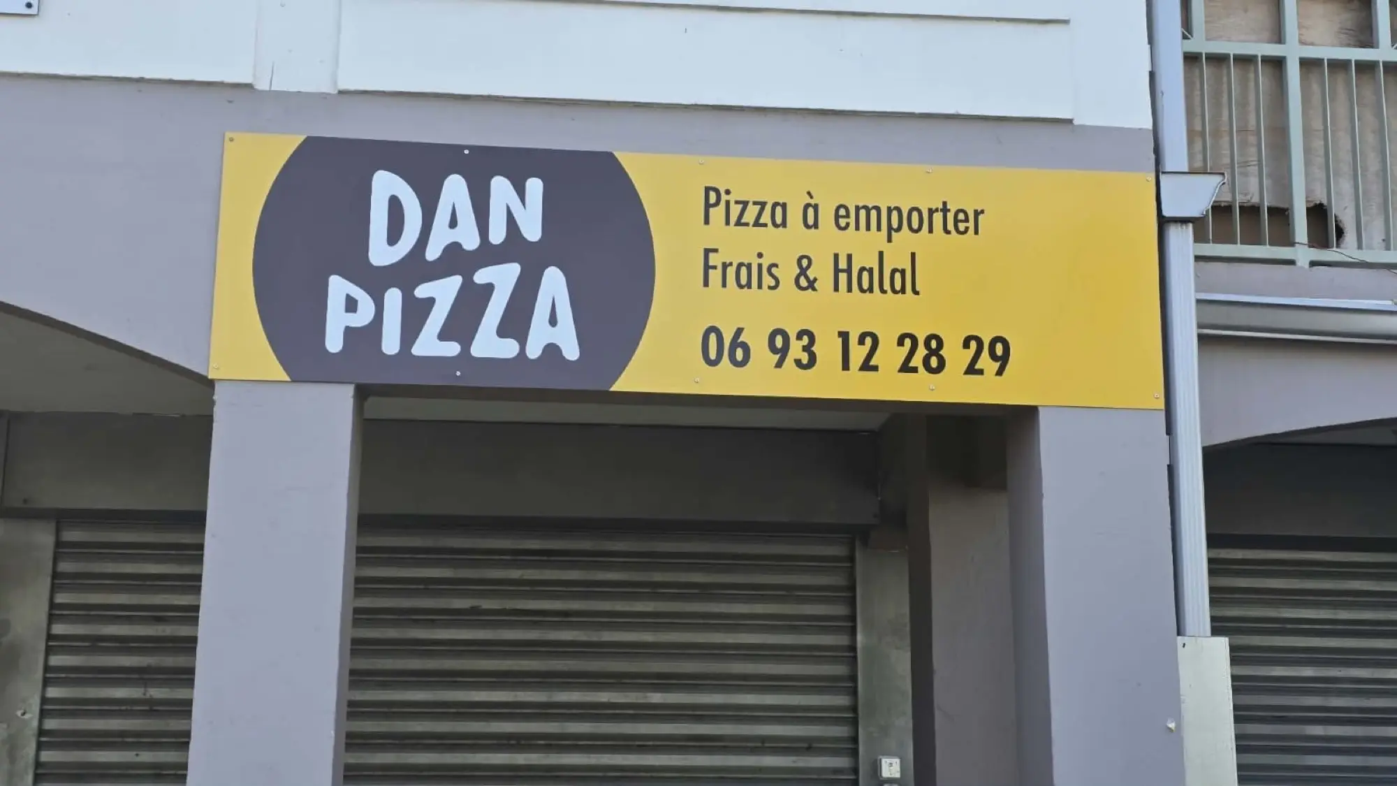 Enseigne bandeau jaune et gris Dan Pizza au-dessus d'une devanture à rideaux métalliques